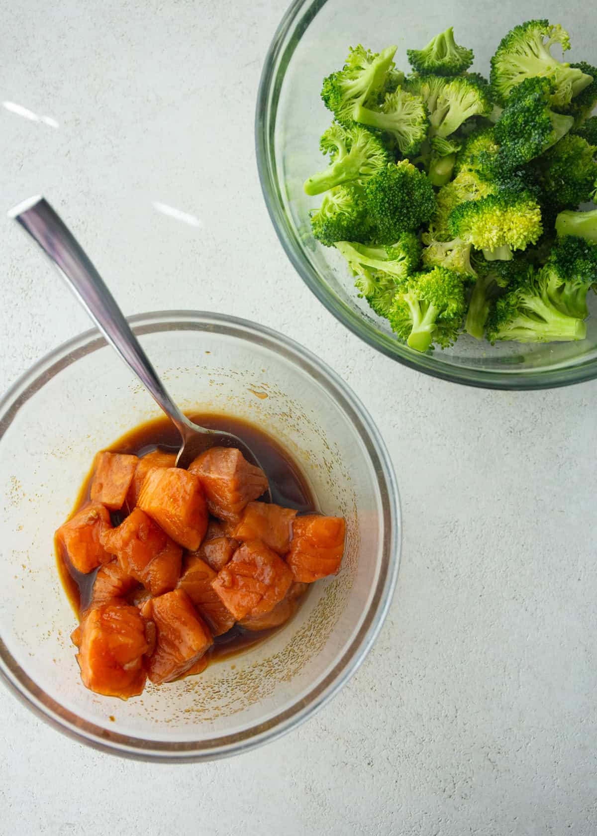 overhead image of salmon marinating and broccoli florets in clear glass bowls