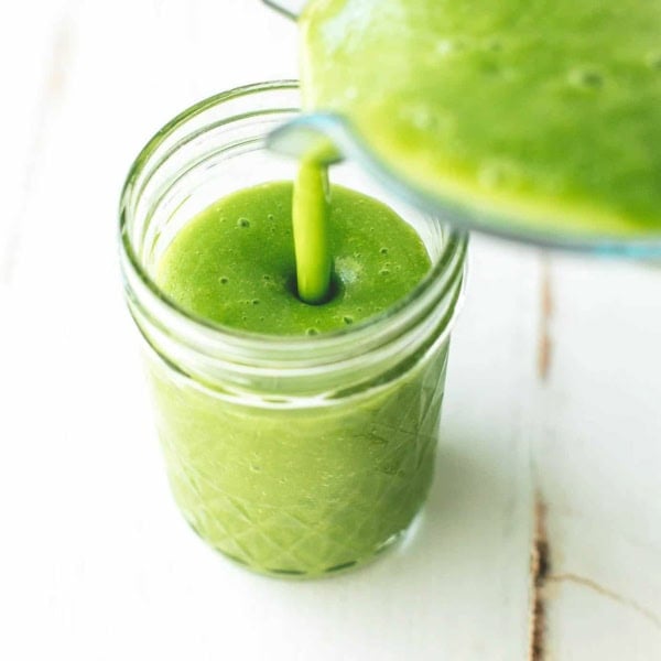 a pitcher of green smoothie pouring into a small mason jar on a white table