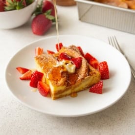 a slice of brioche french toast on a plate, topped with cut strawberries and being drizzled with syrup (square image)