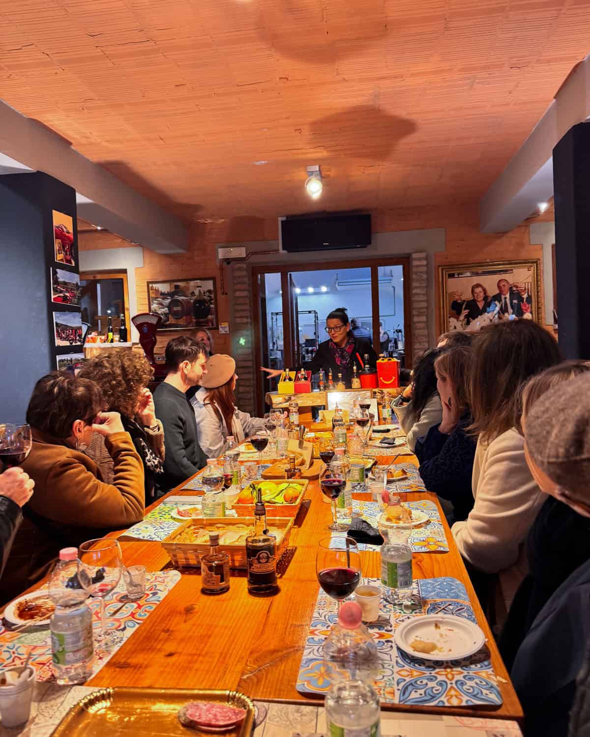 a group of about 20 people sitting around a breakfast table, eating cheese, meat, frittata, and bread with glasses of red wine